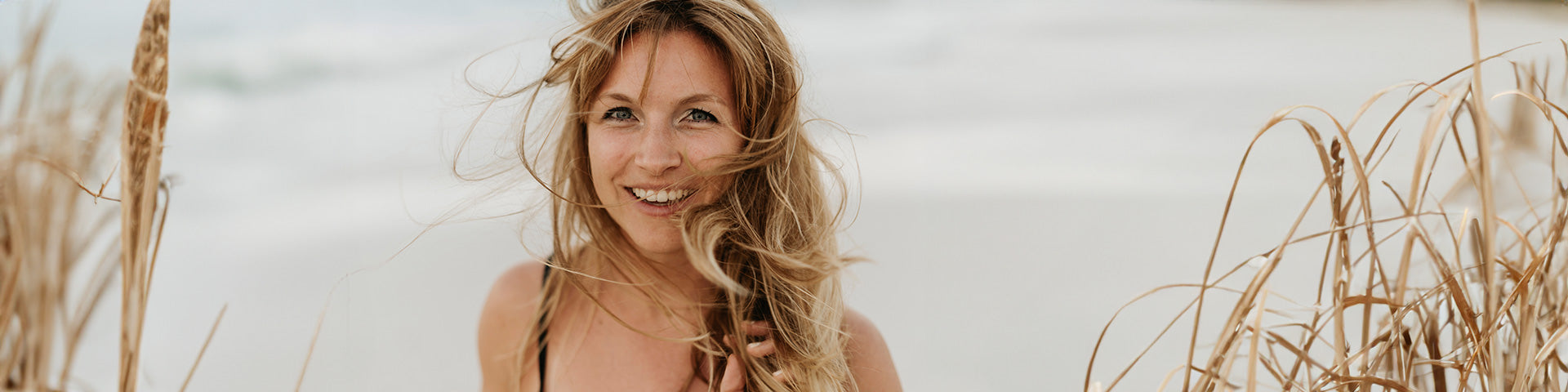 Eine Frau mit vom Wind zerzaustem Haar lächelt in die Kamera, während sie zwischen hohen Strandgräsern an einem Sandstrand steht, während das Meer im Hintergrund verschwimmt.