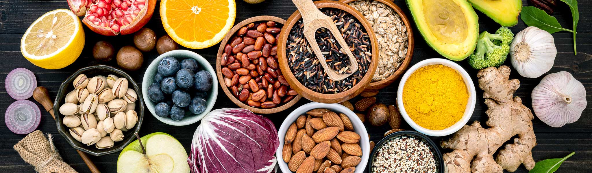Auf einem dunklen Holztisch sind verschiedene Früchte, Gemüse, Nüsse, Samen und Gewürze angeordnet, darunter Orangen, Avocados, Bohnen, Beeren, Mandeln, Knoblauch, Kurkuma, Ingwer und Blattgemüse.