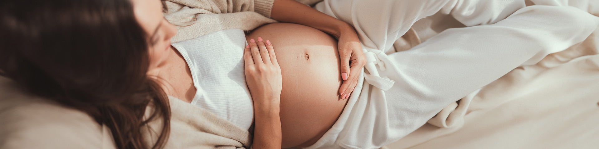 Eine schwangere Frau in bequemer, heller Kleidung liegt auf einem Bett und streichelt mit beiden Händen sanft ihren nackten Babybauch.