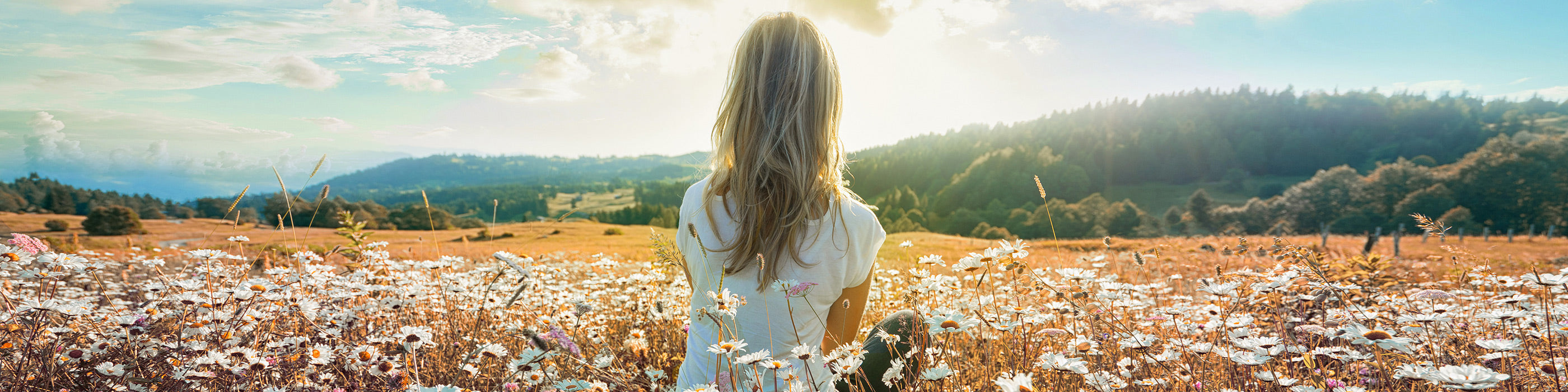 Eine Frau mit langem blondem Haar steht in einem Feld voller Wildblumen und blickt auf einen strahlenden Sonnenaufgang über sanften grünen Hügeln und einem Wald in der Ferne.