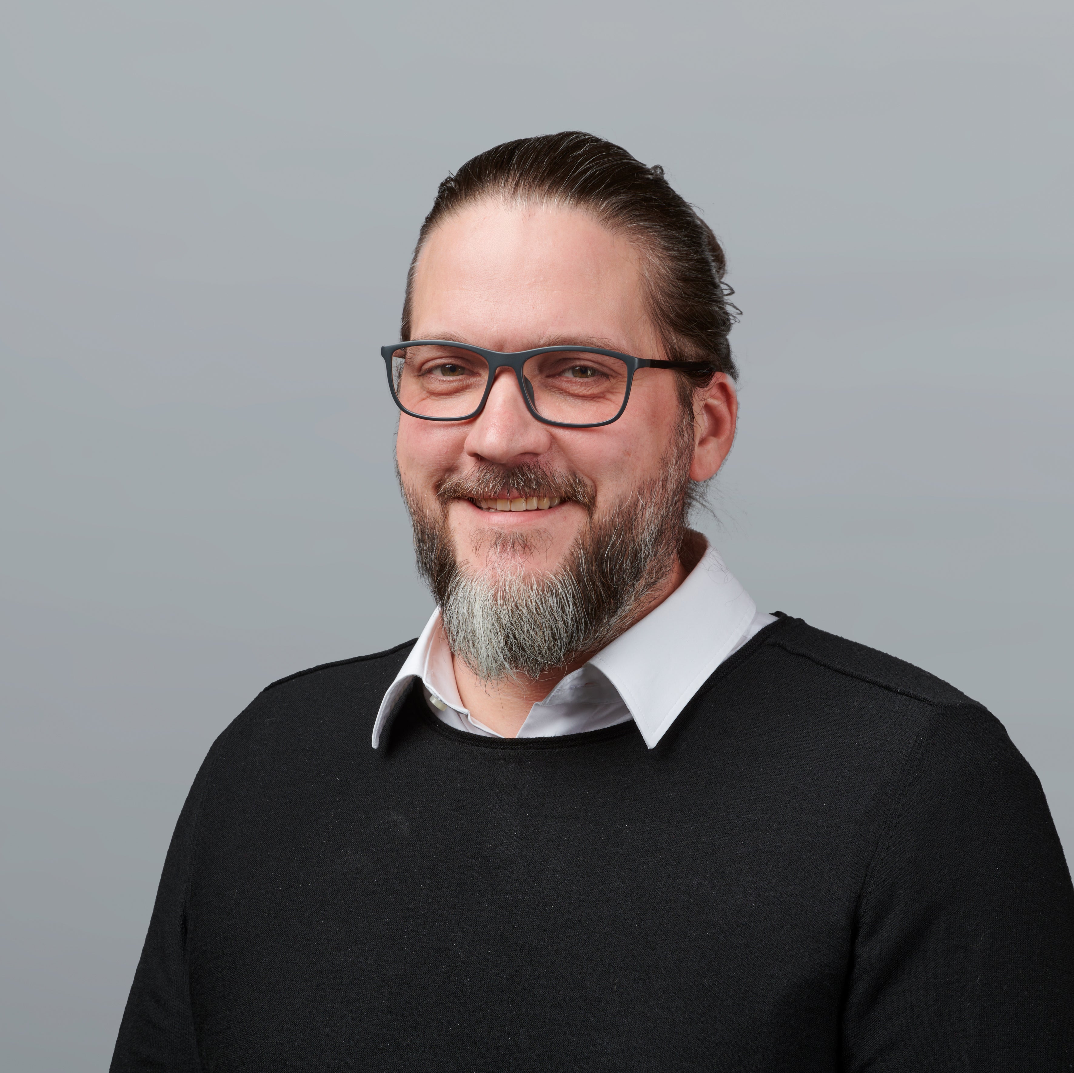 Ein Mann mit Brille, Bart und langen, zurückgebundenen Haaren lächelt. Er trägt einen schwarzen Pullover über einem weißen Hemd mit Kragen und ist vor einem schlichten hellgrauen Hintergrund zu sehen.