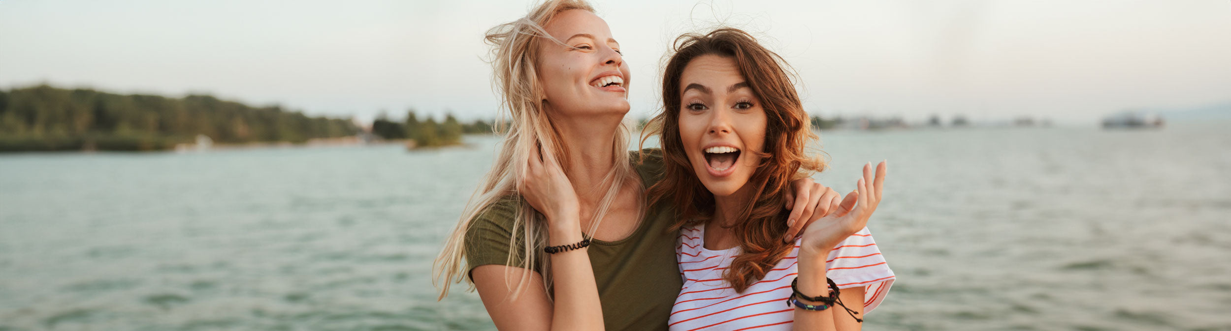 Zwei junge Frauen stehen dicht beieinander am Wasser, lächeln und lachen. Die eine trägt ein grünes Hemd, die andere ein weiß-rot gestreiftes. Im Hintergrund sind ein ruhiger See und Bäume in der Ferne unter einem sanften Himmel zu sehen.